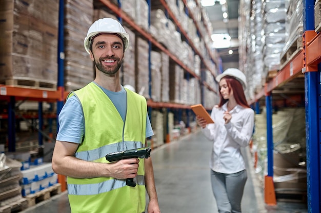 Warehouse worker with a QR scanner and his female colleague with the tablet standing in the aisle with cargo boxes