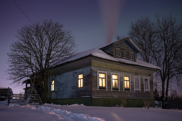 Warm light from windows cozy old russian village house in the bitter cold winter night landscape with snow stars smoke from furnace