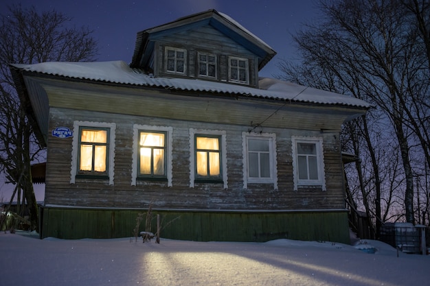 Warm light from windows cozy old russian village house in the bitter cold winter night landscape with snow stars smoke from furnace