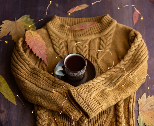 warm sweater sleeves hug a cup of coffee, autumn still life with coffee and yellow leaves and garlands