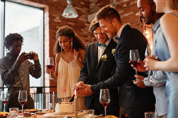 Warm toned portrait of happy gay couple cutting cake together during wedding reception same sex marr