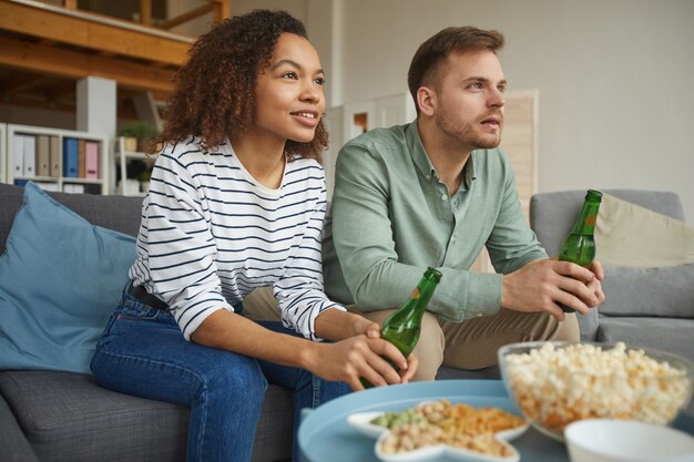 Warm-toned portrait of modern mixed-race couple watching TV at home and drinking beer while sitting on sofa in cozy apartment