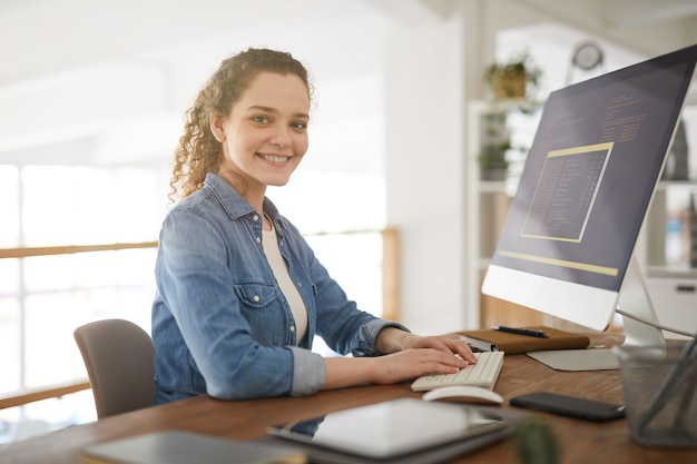 Warm-toned portrait of young woman using computer and smiling at camera while working at desk in software development agency, female IT professional, copy space