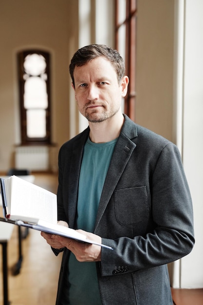 Warm toned vertical portrait of male college professor looking at camera in classroom hall and holdi