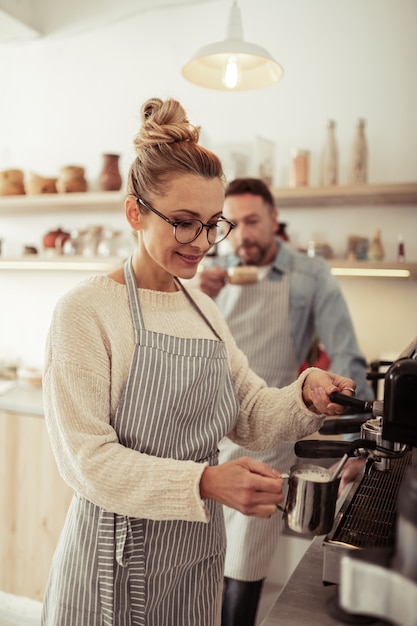 Warming milk. Smiling spectacled barista standing by the coffee machine and frothing milk for cappuccino.