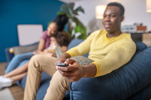 Watch tv. TV remote control in hand of dark-skinned man in yellow sweater sitting relaxed at home on couch wife and daughter behind