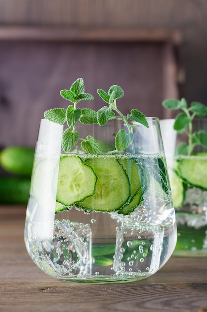 Water with pieces of cucumber, ice and mint leaves in a transparent glass on wooden table