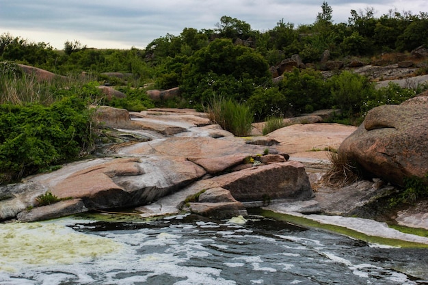 Waterfall flows among high rocks, red stones and cliffs, green bushes and trees