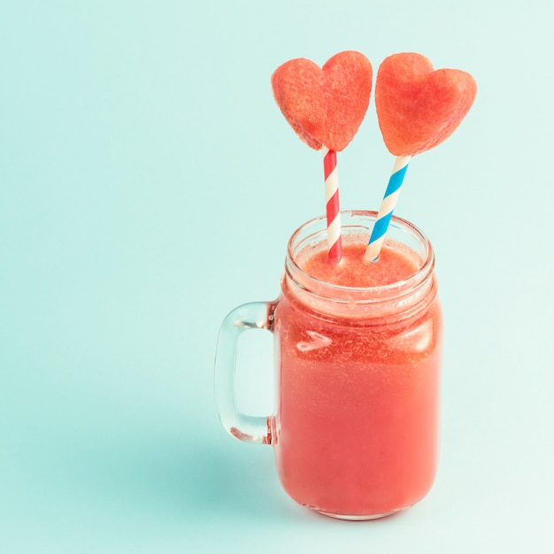 Watermelon smoothie with heart-shaped slices on straws in a glass jar on blue background 