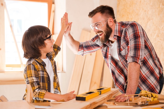 We did it! Cheerful young male carpenter and his son giving high-five to each other while working in workshop