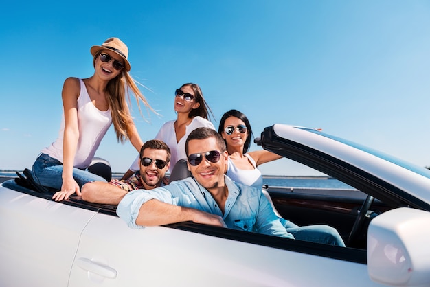 We love spending time together! Group of young happy people enjoying road trip in their white convertible and smiling at camera