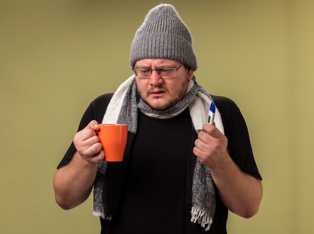 Weak middle-aged ill male wearing winter hat and scarf holding cup of tea with thermometer 