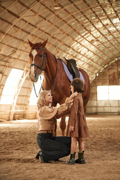 Wearing the jockey clothes Woman with her little daughter is with horse indoors