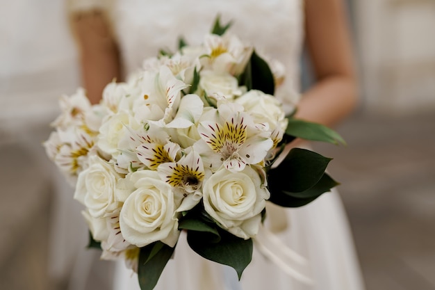 Wedding bouquet closeup with white roses and green leaves. Bride in dress holds bouquet.