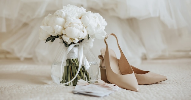 Wedding bouquet of peonies flowers in a vase stands on the bed of the newlyweds with invitations and shoes on the background