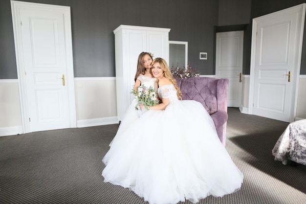 Wedding Bride in beautiful dress sitting on sofa indoors in white studio interior like at home Trendy wedding style shot in full length Young attractive caucasian brunette model like a bride