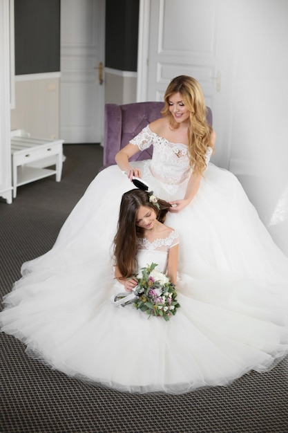 Wedding Bride in beautiful dress sitting on sofa indoors in white studio interior like at home Trendy wedding style shot in full length Young attractive caucasian brunette model like a bride