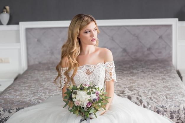 Wedding Bride in beautiful dress sitting on sofa indoors in white studio interior like at home Trendy wedding style shot in full length Young attractive caucasian brunette model like a bride