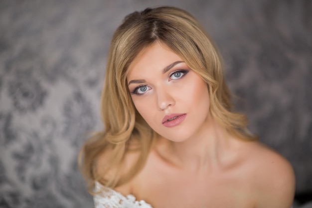 Wedding Bride in beautiful dress sitting on sofa indoors in white studio interior like at home Trendy wedding style shot in full length Young attractive caucasian brunette model like a bride