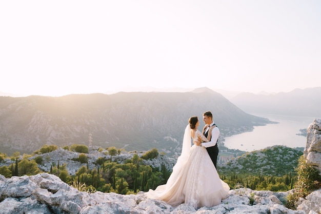 A wedding couple stands on top of a mountain with panoramic views of the bay of kotor at sunset the