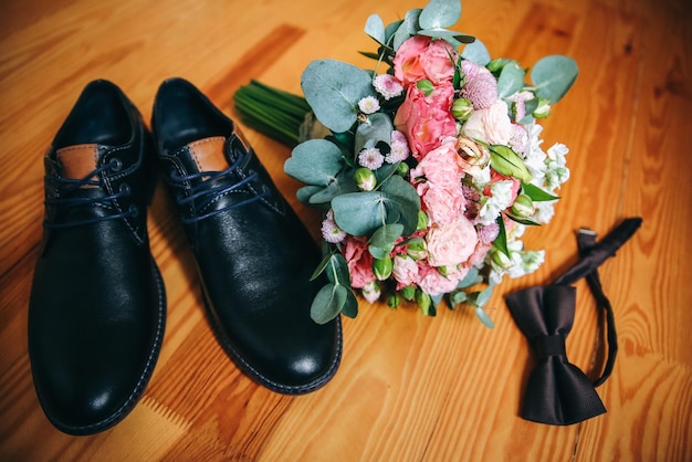 Wedding day accessories Groom's shoes with bouquet on the wooden background Golden rings and bow tie are nearby Stylish things