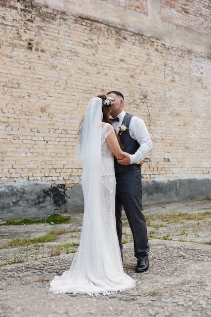 Wedding day bride in long white dress ang groom hugging and kissing outdoors near brick wall coupl