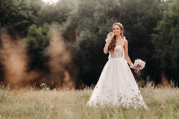 On the wedding day, an Elegant bride in a white long dress and gloves with a bouquet in her hands stands in a clearing enjoying nature