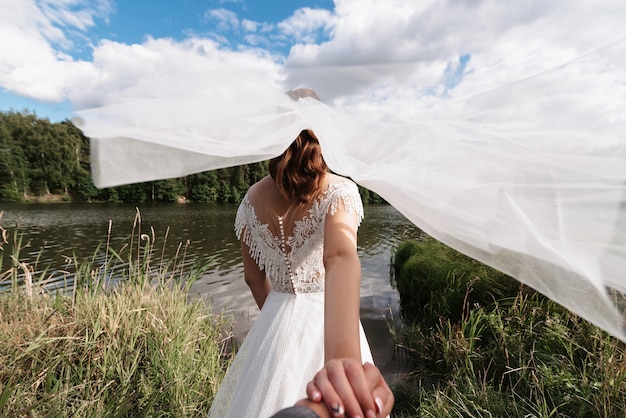 Wedding follow me concept. The bride holds the hand of the groom on the wedding day in the summer in nature by the river