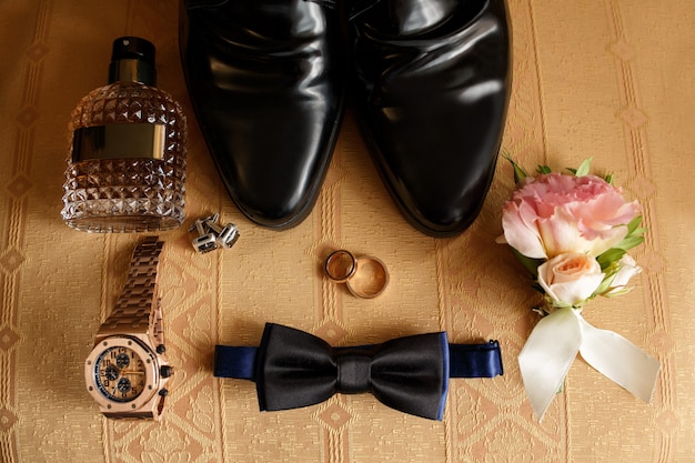 Photo wedding groom accessories on chair: black shoes, wrist watch, perfume bottle, bow tie near wedding rings