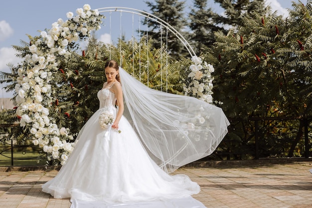 Wedding photography in nature A brunette bride in a veil and a tiara in her hair poses near a white arch made of flowers in the shape of a circle The veil is in the air Sunny day Wedding ceremony