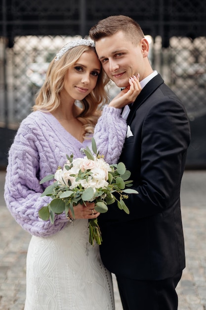 Wedding portrait of blonde bride with curly hair wearing wedding dress, lilac jacket, having bandeau on head and bouquet and bridegroom in black suit standing close and looking at viewer outdoors