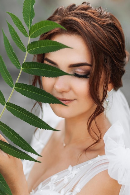 Wedding portrait The bride in a white dress is posing holding a leaf near her face and looking into the lens Nice makeup Portrait of the bride