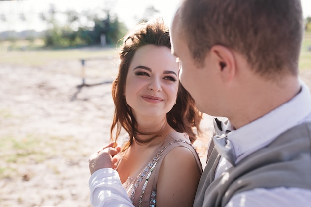 Wedding at sunset. Couple is sitting on a bench under a tree. Beige dress with sparkles. Light suit with a bow tie.