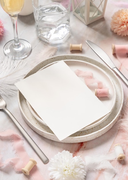 Wedding Table place with a blank card near flowers, silk ribbons and feathers, closeup. Romantic scene with vertical paper card mockup on a plate on a marble table