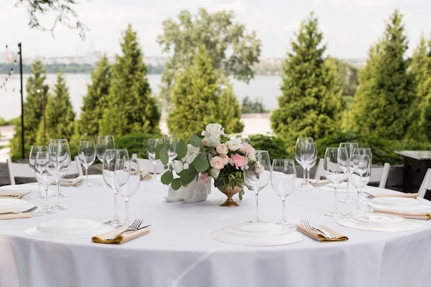 Wedding table setting decorated with fresh flowers in a brass vase. Wedding floristry. Banquet table for guests outdoors with a view of green nature. Bouquet with roses, eustoma and eucalyptus leaves