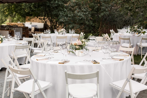 Wedding table setting decorated with fresh flowers in a brass vase. Wedding floristry. Banquet table for guests outdoors with a view of green nature. Bouquet with roses, eustoma and eucalyptus leaves