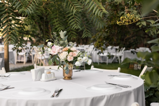 Wedding table setting decorated with fresh flowers in a brass vase. Wedding floristry. Banquet table for guests outdoors with a view of green nature. Bouquet with roses, eustoma and eucalyptus leaves