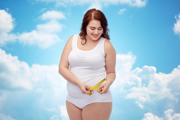 weight loss, diet, slimming, size and people concept - happy young plus size woman in underwear measuring tape over blue sky and clouds background