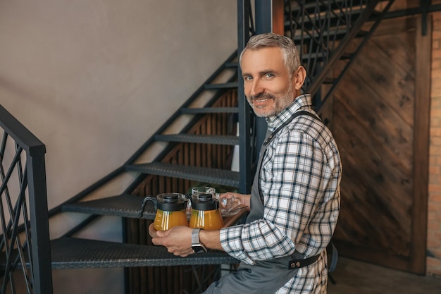 Welcome. Happy man in apron holding tray of fresh juice climbing stairs turning smiling at camera in cafe