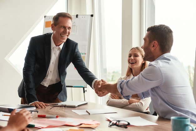 Welcome two cheerful colleagues shaking hands and smiling while sitting in the modern office