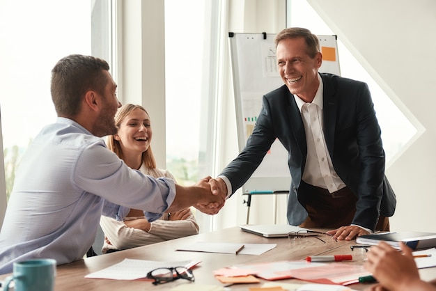 Welcome two cheerful colleagues shaking hands and smiling while sitting in the modern office