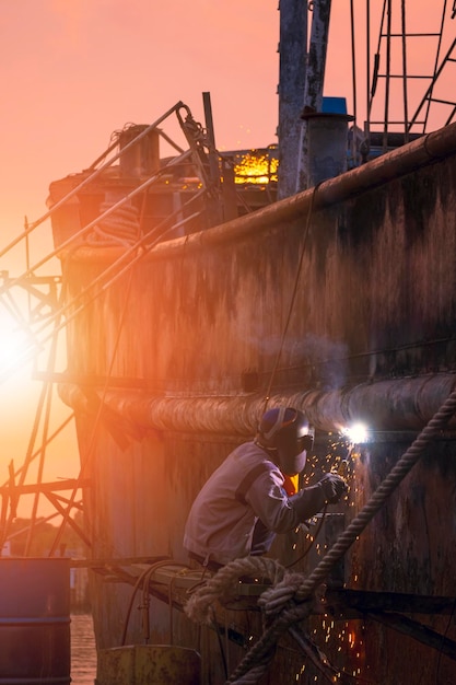 Welder on platform is welding metal wall of the old fishing vessel in shipyard area at sunset time