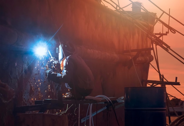 Welder on scaffold is welding the old vessel hull on overtime in shipyard area at sunset time