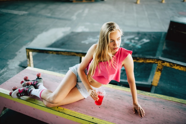 Well-built and slim girl is posing. She looks like a model. Girl is holding red juice in right hand. Also she wears rollers.