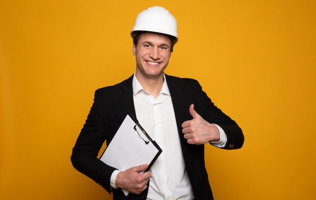 Well done! Young attractive man in formal clothes and a hard hat is looking in the camera, showing thumbs up and holding his work notes.