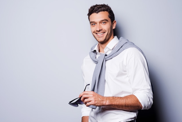 Well-dressed handsome. Handsome young man in smart casual wear holding his sunglasses and smiling while standing against grey background