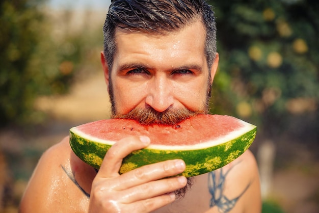 Wet tanned bearded man with a big red mustache laughs and holds a piece of ripe red watermelon in the rain in a green garden