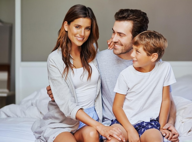 What are our plans for the day Portrait of a cheerful young family seated on bed while holding each other and looking at the camera at home in the morning