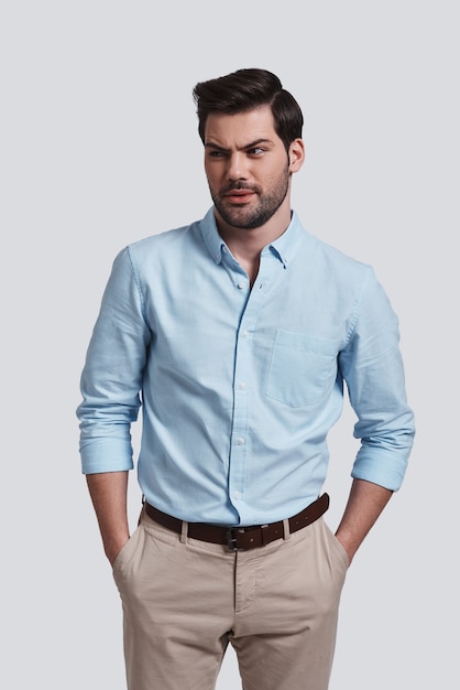 What?  Confused young man looking away and keeping hands in pockets while standing against grey background