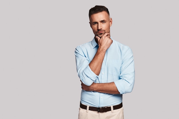 What decision to make? Handsome young man keeping hand on chin and looking at camera while standing against grey background